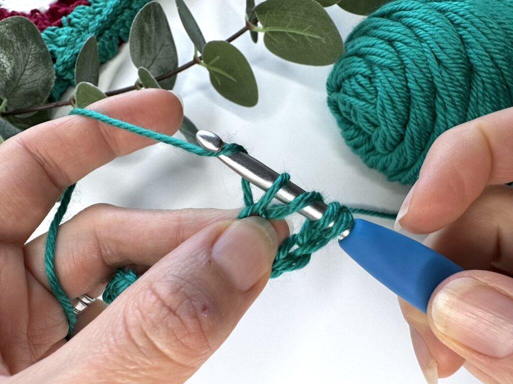 Hands doing crochet stitch with green yarn and a blue hook