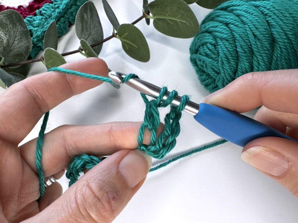 2 hands hold green crochet and a hook and are wrapping the yarn around the hook to create a stitch 
