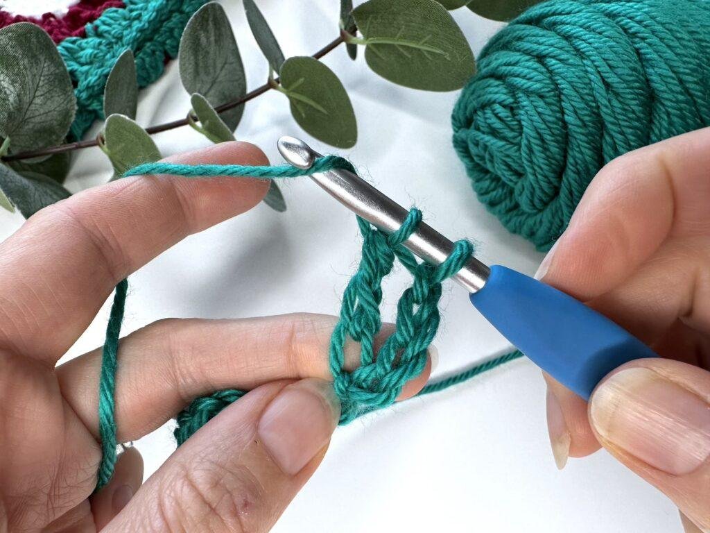 with 2 loops on a crochet hook, 2 hands are wrapping the yarn around the hook ready to complete a crochet stitch in some crochet fabric