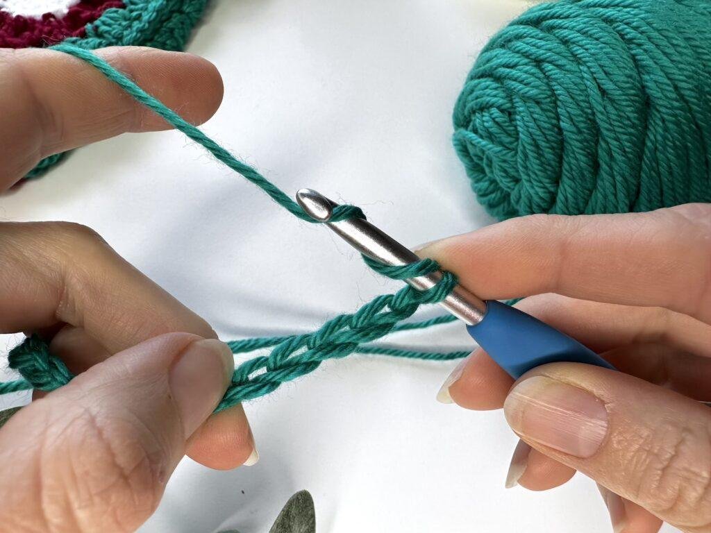2 hands hold a green crochet chain and a crochet hook and are wrapping the yarn around the hook