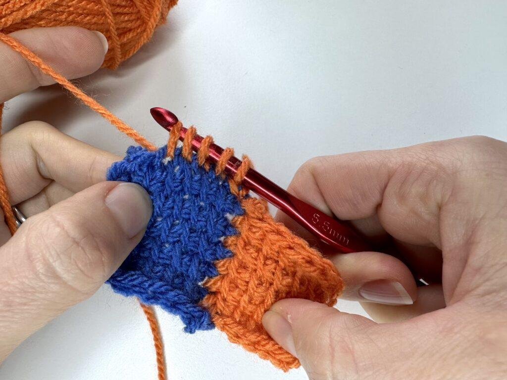 Against a white background, 2 hands are crocheting using orange yarn and a red hook. They are pulling up loops onto the hook