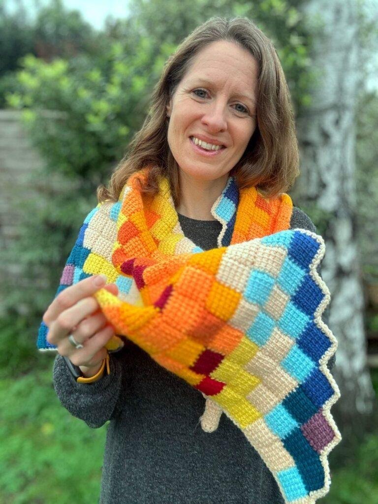 A lady wears a colourful, triangle crochet shawl and holds it up to show the camera, so we can see the different coloured entrelac crochet blocks