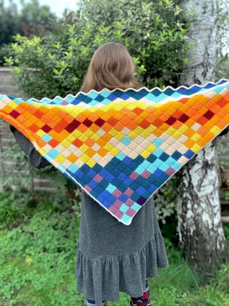 A woman stands in a garden with her back to the camera holding out a crochet triangle shawl made out of lots of coloured squares