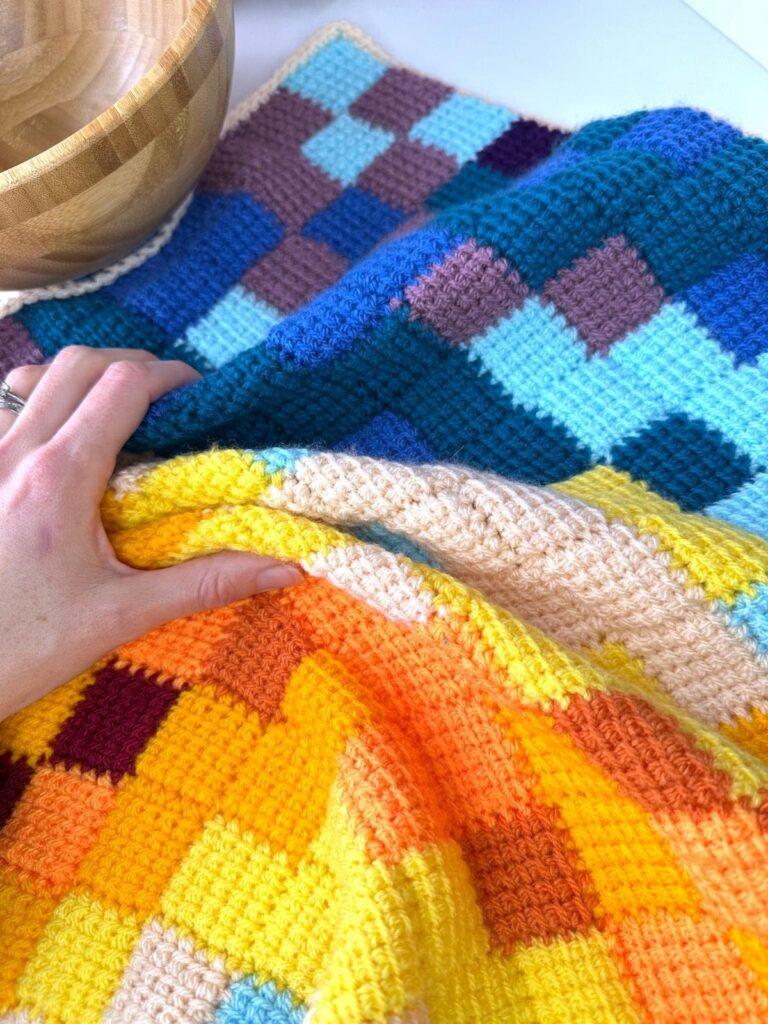 A crochet block shawl is on a table with a wooden bowl. A hand is scrunching up the shawl and we can see lots of different coloured squares on it