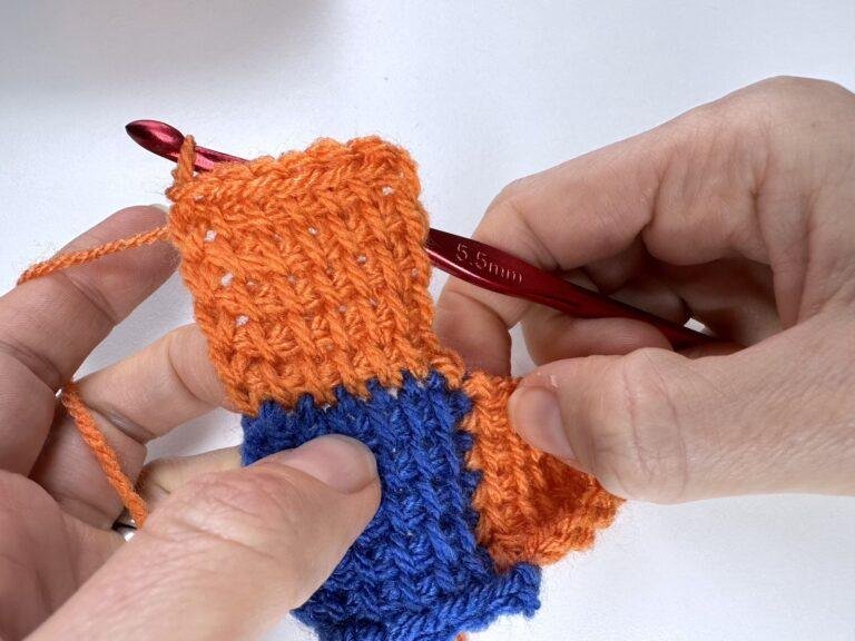 A completed Tunisian crochet entrelac final square in orange is joined to a blue and an orange square and held in 2 hands.
