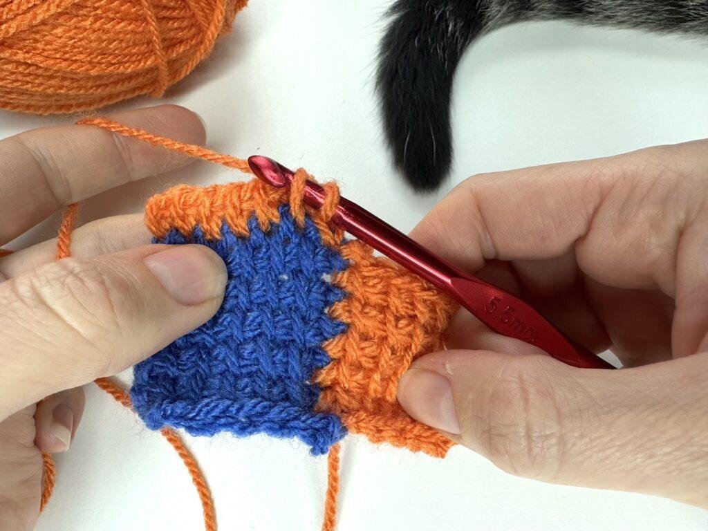 A forward pass is being completed in Tunisian crochet entrelac with 2 hands, orange yarn and a red crochet hook