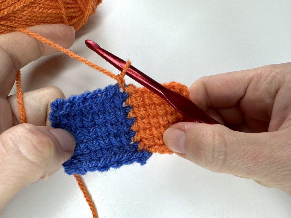 2 hands are holding 2 squares of tunisian crochet entrelac and have jut joined on orange yarn for the next square