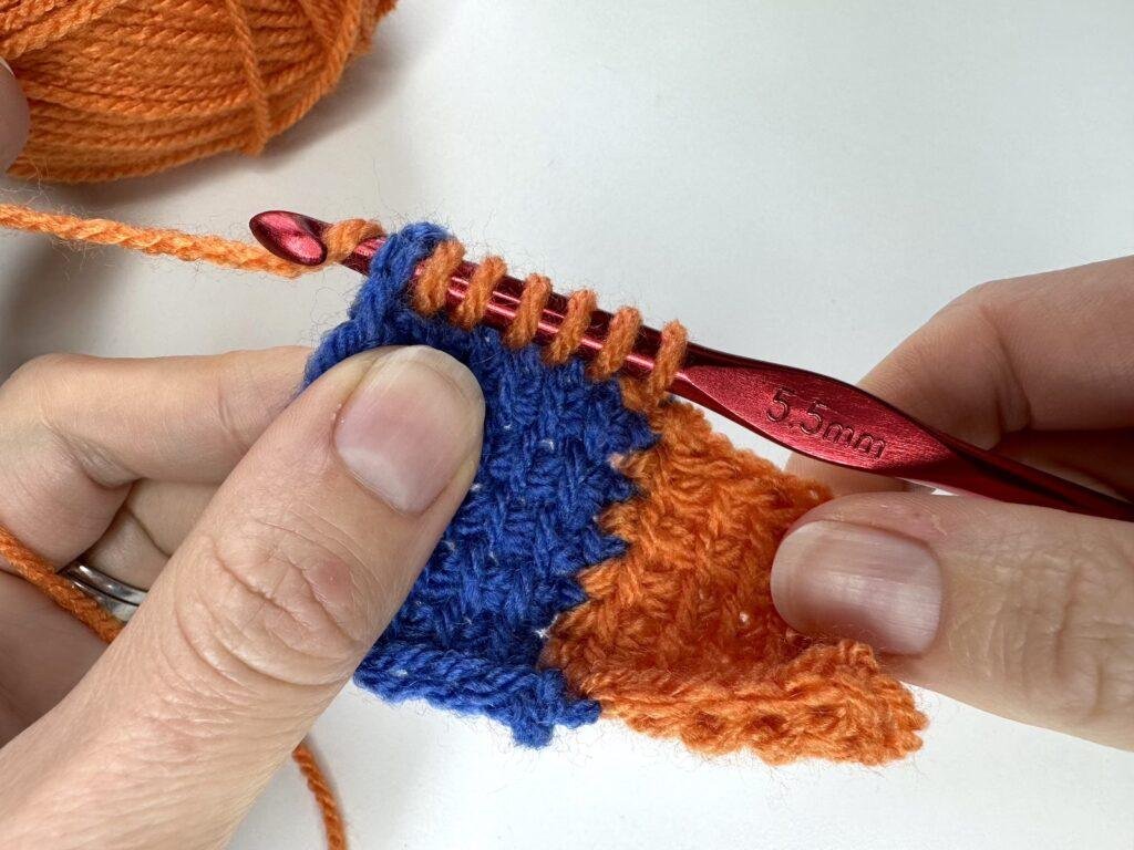 In Tunisian crochet entrelac, a final square is being made by 2 hands and a red hook and orange yarn. The hook is pulling up loops and has just reached the last stitch