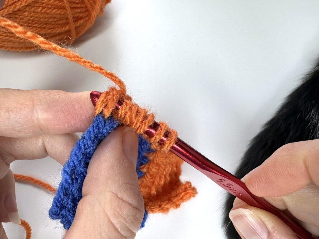 The last stitch of the final square has been reached by 2 hands and a red crochet hook in Tunisian crochet entrelac.