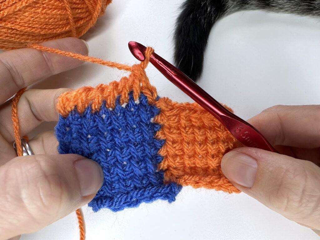 A Tunisian crochet entrelac final square held in 2 hands. 1 row of a third square has just been completed in orange yarn