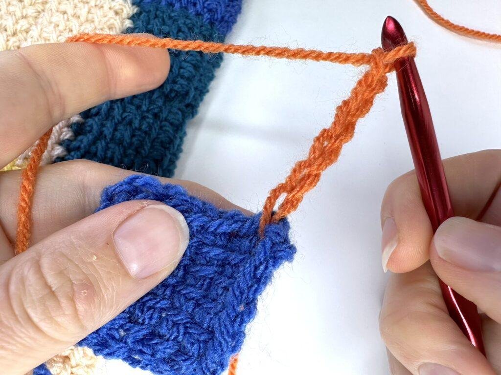 2 hands are holding a blue crochet square and have just joined orange yarn and made a chain with a red crochet hook in tunisian crochet entrelac 