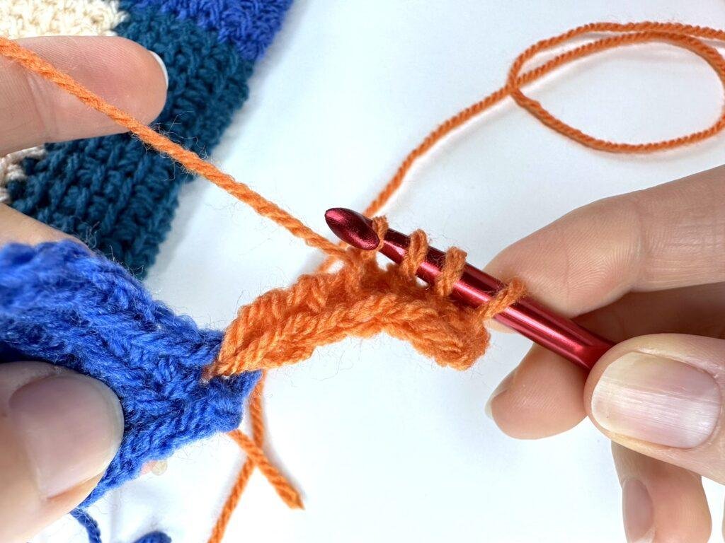 Tunisian crochet entrelac initial square forward pass is being done by a red crochet hook and orange yarn. It is being joined to a blue square already made.