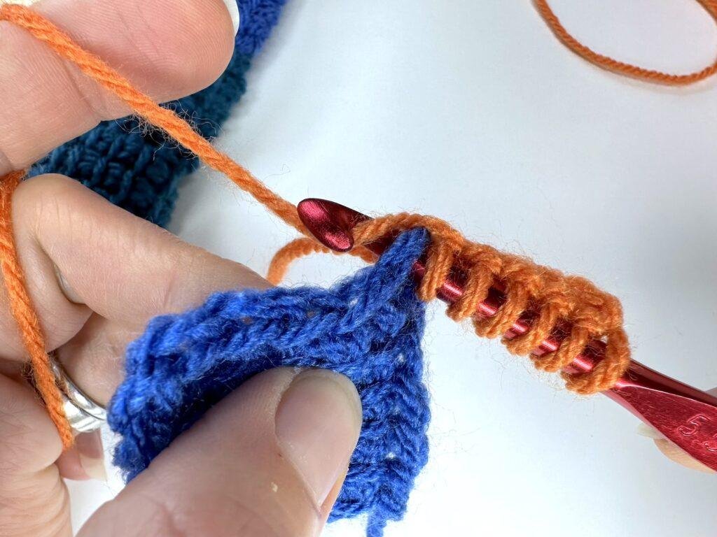 A hand holds a blue tunisian crochet entrelac first square and is joining on an initial square in orange yarn