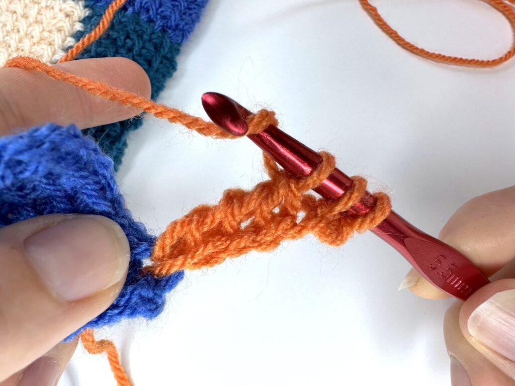 2 hands are partway through a return pass in Tunisian crochet entrelac making an initial square in orange yarn