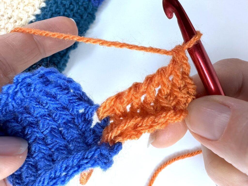 2 rows of the Tunisian crochet entrelac initial square have been completed in orange yarn, joining to a blue square, both held in 2 hands.
