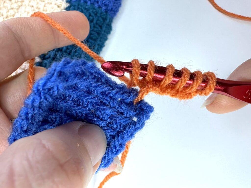 2 hands are showing the first row of a Tunisian crochet entrelac initial square using orange yarn and joining it to a blue first square