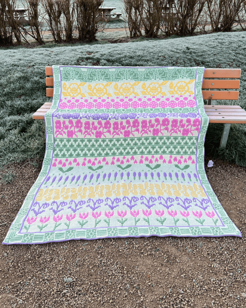 A large crochet blanket a draped on a bench outside and it features pastel coloured flowers in strips