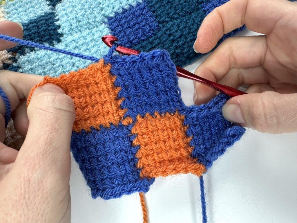 2 hands are holding a blue and orange sample of Tunisian crochet entrelac showing a completed joining square 