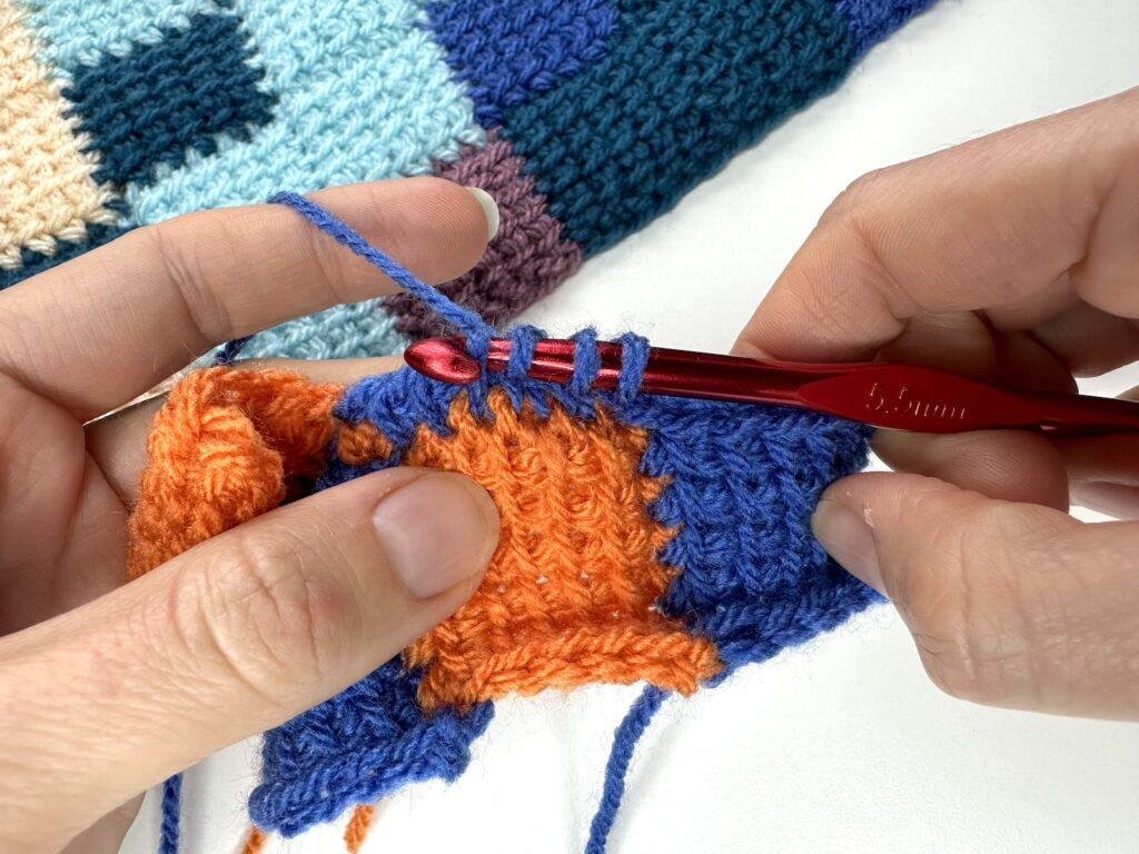 2 hands are gripping a sample of orange and blue crochet squares and are working on joining on another blue square by pulling up loops on to the hook