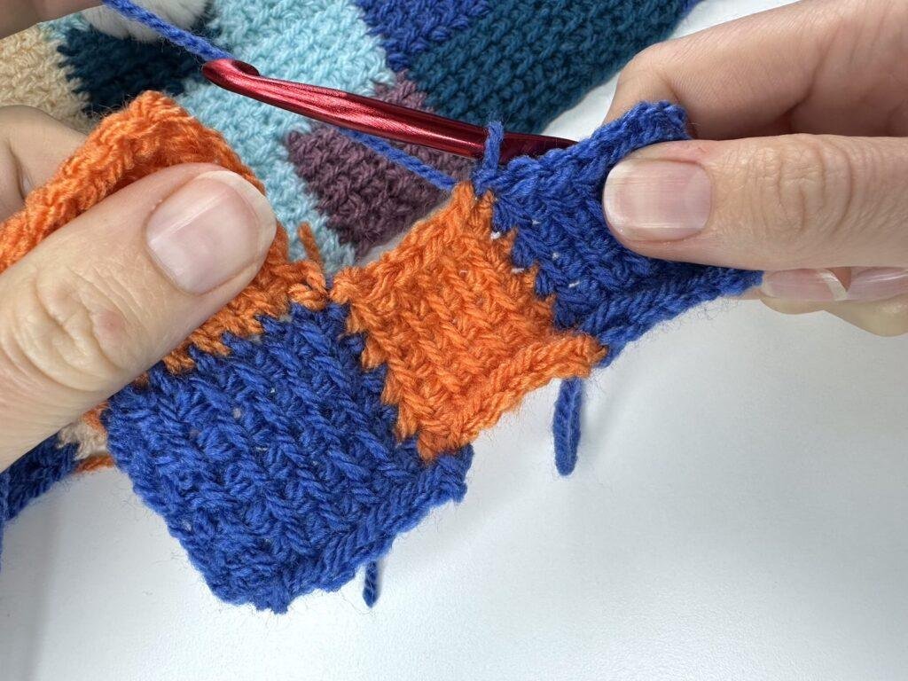 A person is holding a crochet sample of 2 orange and 2 blue squares joined together and has just joined blue yarn for the Tunisian crochet entrelac joining square