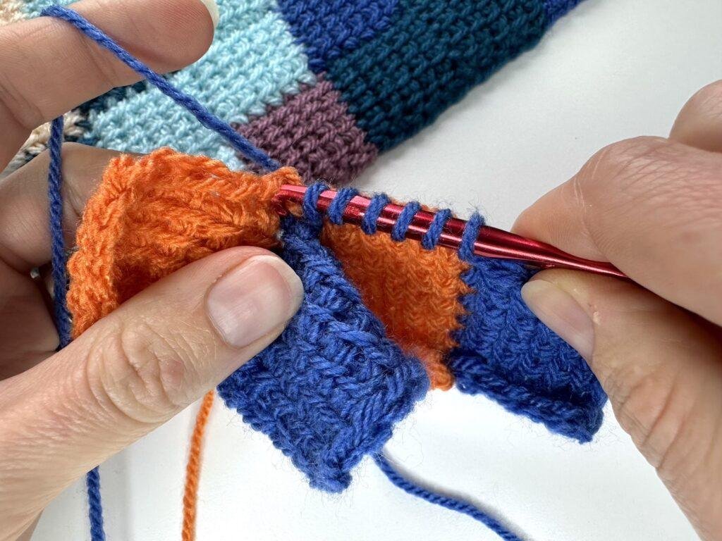 The last stitch is being worked into the side of the next orange crocheted square in Tunisian crochet entrelac. 2 hands hold the work and a red crochet hook