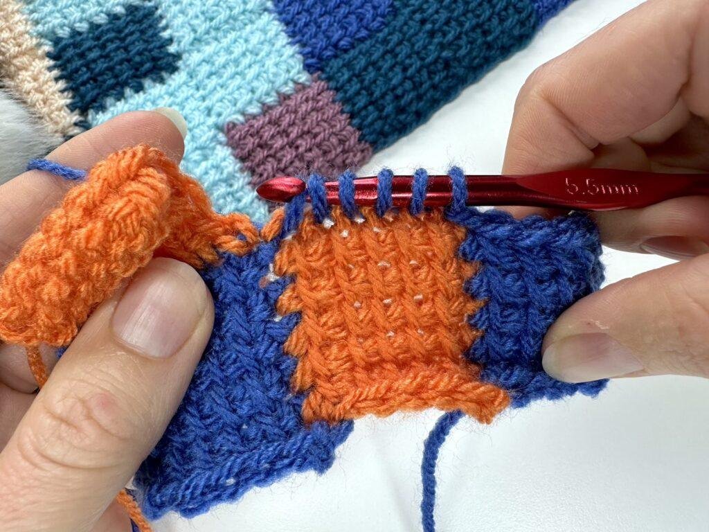 2 hands are partway through pulling up loops with a red crochet hook in Tunisian crochet entrelac joining blue yarn onto an orange square which is joined to 2 blues squares
