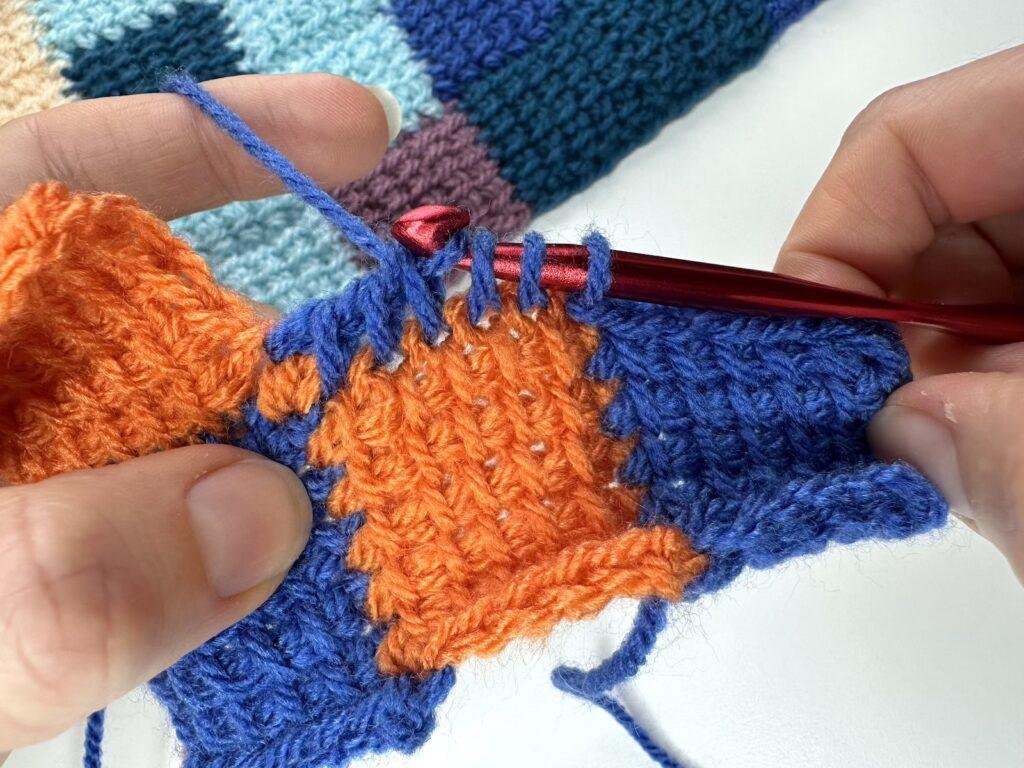 2 hands holding yarn, hook and a part made crochet entrelac sample are making a return pass