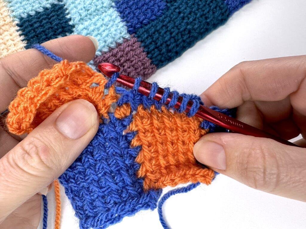 2 hands are doing Tunisian crochet entrelac and making a joining square. They have just reached the last stitch and are pulling up a loop in blue yarn working into an orange square of crochet