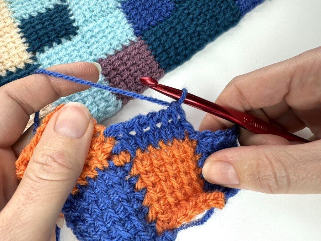 2 hands are holding a crochet Tunisian entrelac sample that is being worked on. It is orange and blue squares and a blue square in the middle of 2 other squares is part complete
