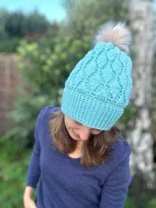 A woman tipping her head forward to show off a pale turquoise coloured crochet beanie with leaf shaped motifs in the texture and natural coloured pom pom. She stands in a garden