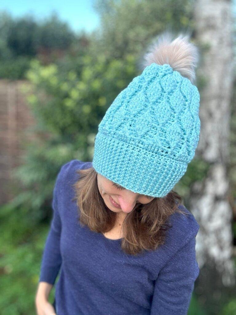 A woman tipping her head forward to show off a pale turquoise coloured crochet beanie with leaf shaped motifs in the texture and natural coloured pom pom. She stands in a garden