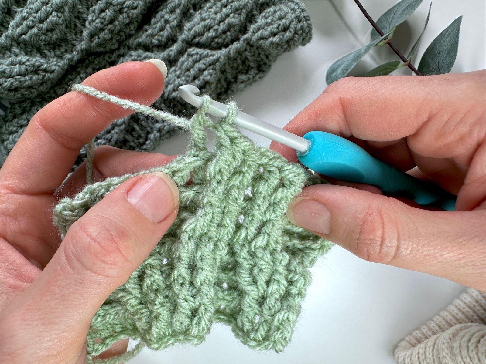 2 hands are partway through crocheting a back post double crochet 2 together with green yarn