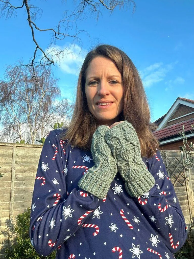 A lady wearing a Christmas dress and green crochet mittens stands in a garden with her hands together held under her chin. The crochet mittens have textured leaves running up and down them