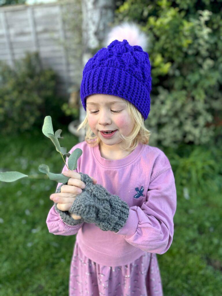 A small child stands in a garden wearing a pink sweater dress, a purple crochet hat with pink Pom Pom and grey crochet fingerless mittens. The hat and mittens have the same leaves motif crocheted into the texture. The girl is holding some leaves and looking at them