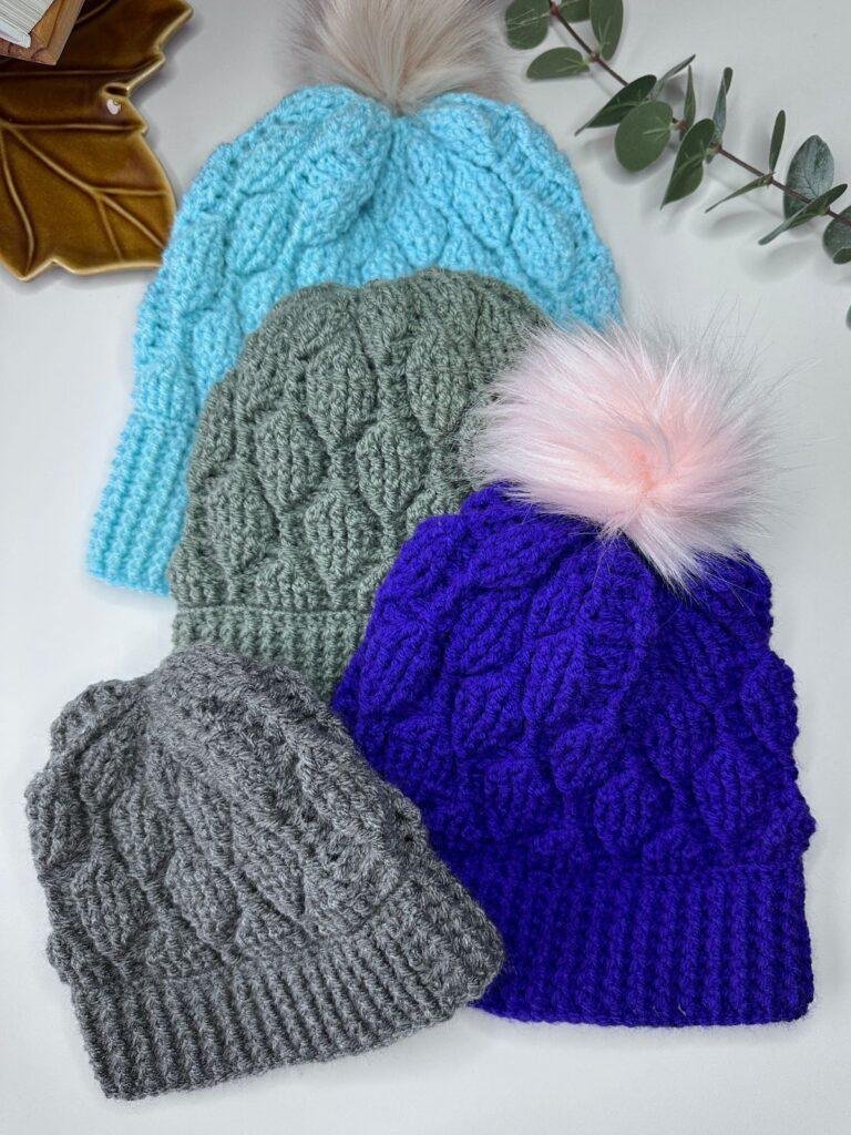 4 crochet hats are on a white surface with a dish and leaves just coming into the shot. The crochet hats are grey, purple, green and blue and 2 have pom poms. They feature textured leaf pattern running down the hats