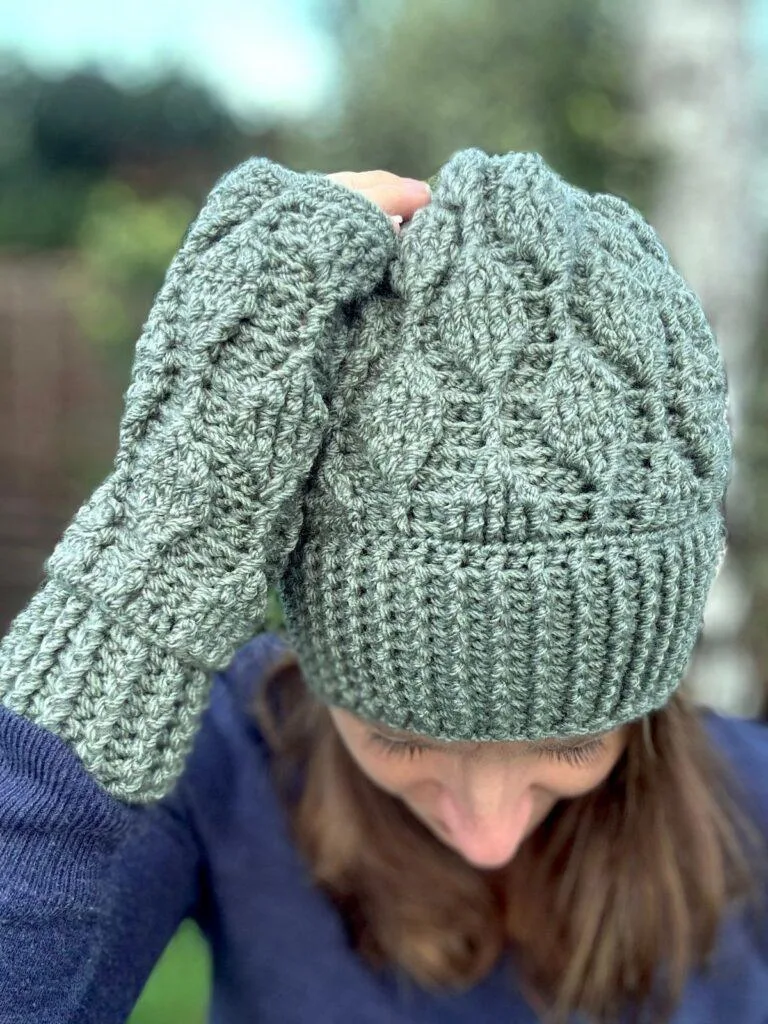 A woman bends her head forwards to the camera and puts her hand on her head. She is wearing a matching crochet fingerless mitten and beanie which are both green and have trailing leaves texture on them