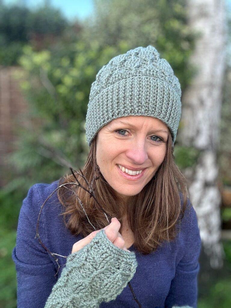 A lady stands in a garden smiling and holding her hair. She is wearing a blue sweater and a green crochet mitten and a matching green crochet hat with deep ribbing and a leaf texture motif all over it