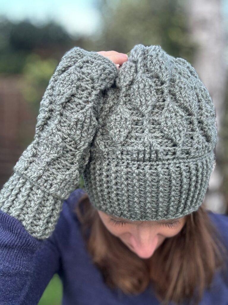 Close up of a woman's head with a green textured crochet hat on. One hand wearing a matching crochet mitten is held to the head. The mitten and hat feature leaf shaped motifs in textured crochet. The woman has long brown hair and a blue sweater