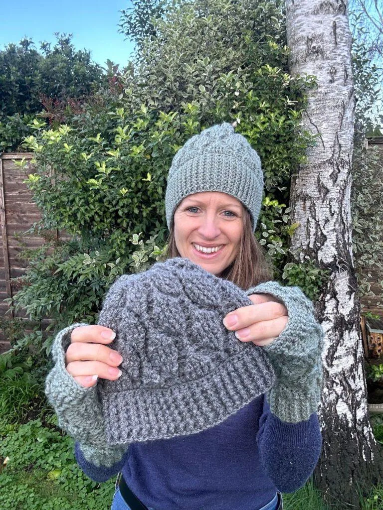 A smiling lady stands in a garden wearing green crochet hat and mittens and holding out a grey crochet hat in front of her. They all have the same textured crochet leaf pattern.