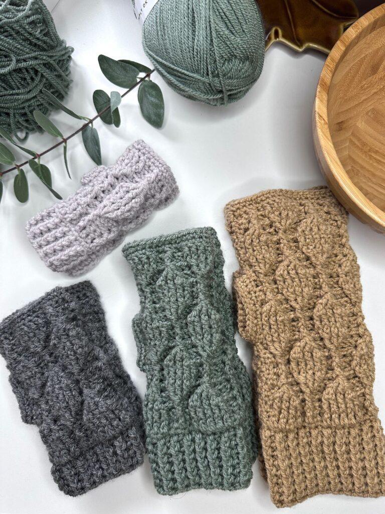 4 pairs of crochet fingerless mittens are laid out on a white surface. They all feature the same leaf motif but are different sizes and colours, light grey, dark grey, green and beige. Around them are yarn, a leaf sprig and a wooden bowl