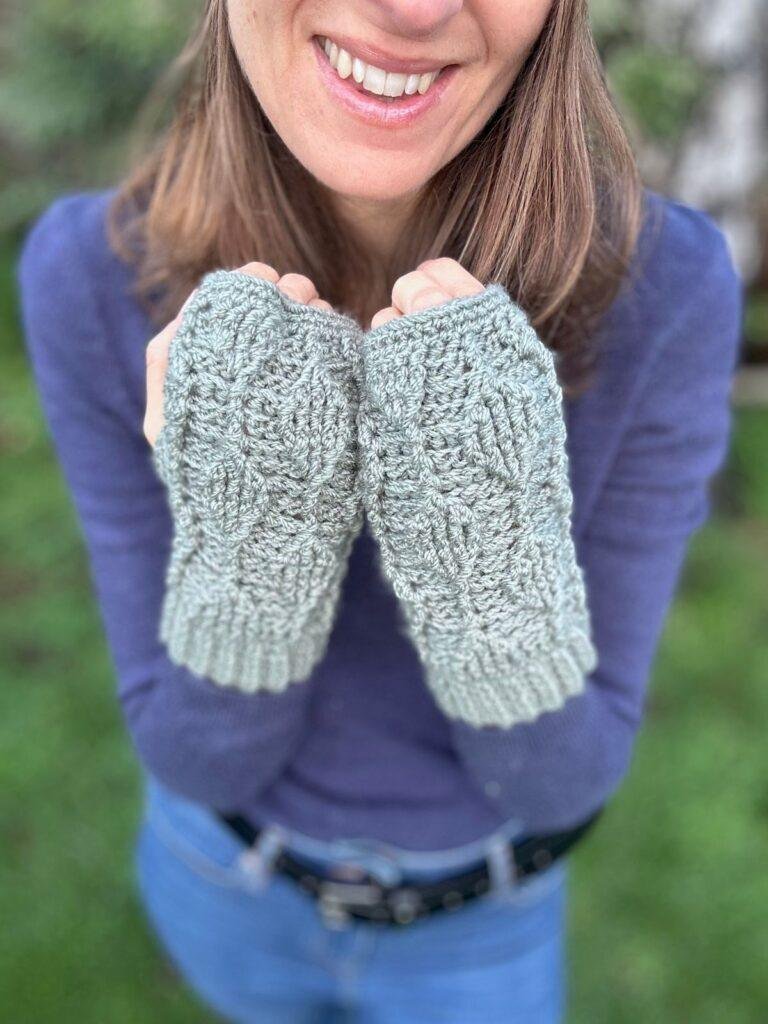 Close up of a lady holding up her hands with crochet fingerless mittens on in front of her. The mittens are green with leaves on them in textured stitch and there is greenery behind