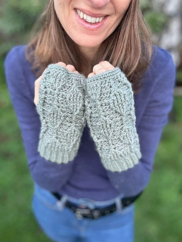 Close up of a lady holding up her hands with crochet fingerless mittens on in front of her. The mittens are green with leaves on them in textured stitch and there is greenery behind