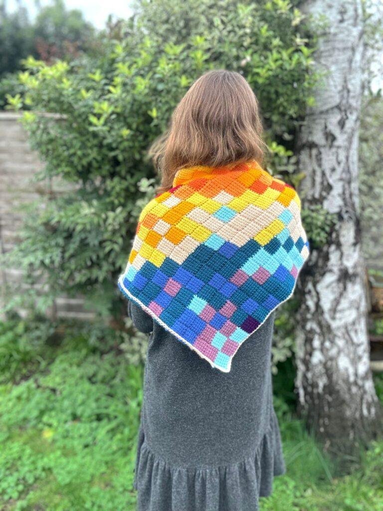 A woman stands in a garden with her back to the camera wearing a grey dress and an entrelac crochet shawl, featuring different coloured squares. 