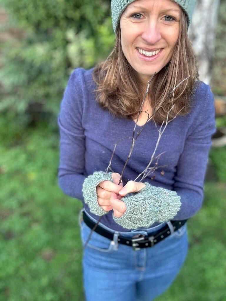 A smiling lady stands in a garden with blue sweater and jeans on wearing green crochet fingerless mittens and a matching hat that is mostly out of shot. She holds a twig in her hands