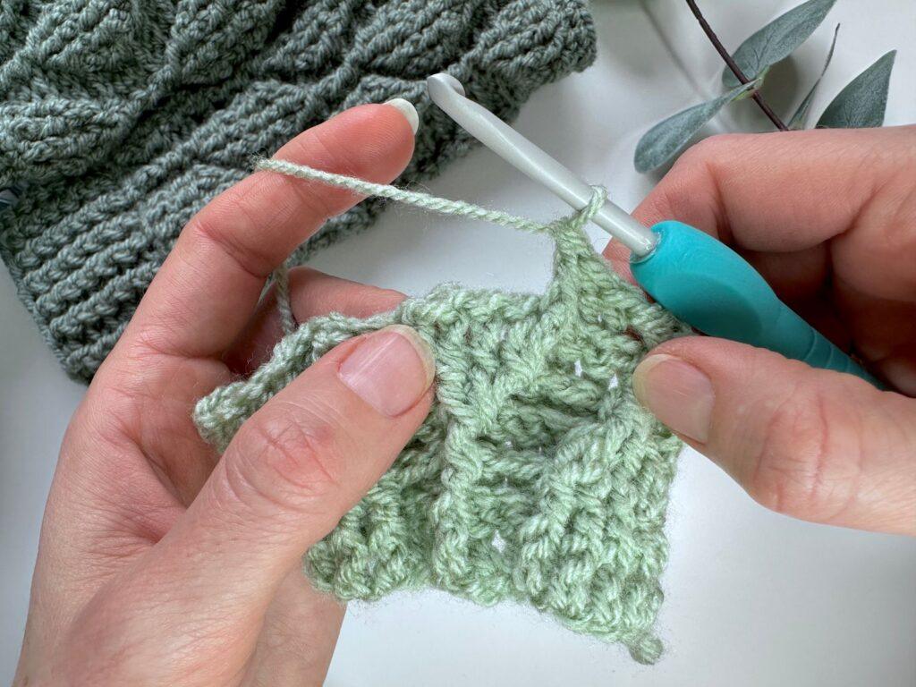 A sample of green crochet held in 2 hands and a crochet hook showing a front post double crochet stitch