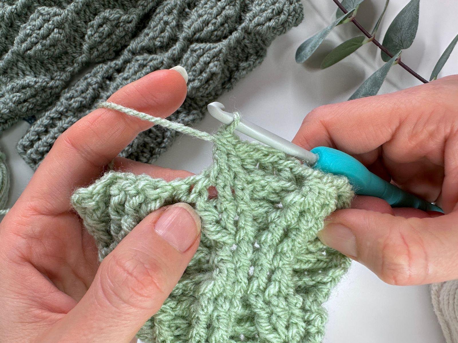 The stitch tutorial continues using the green yarn and 2 hands holding a sample of front and back post double crochet stitches.