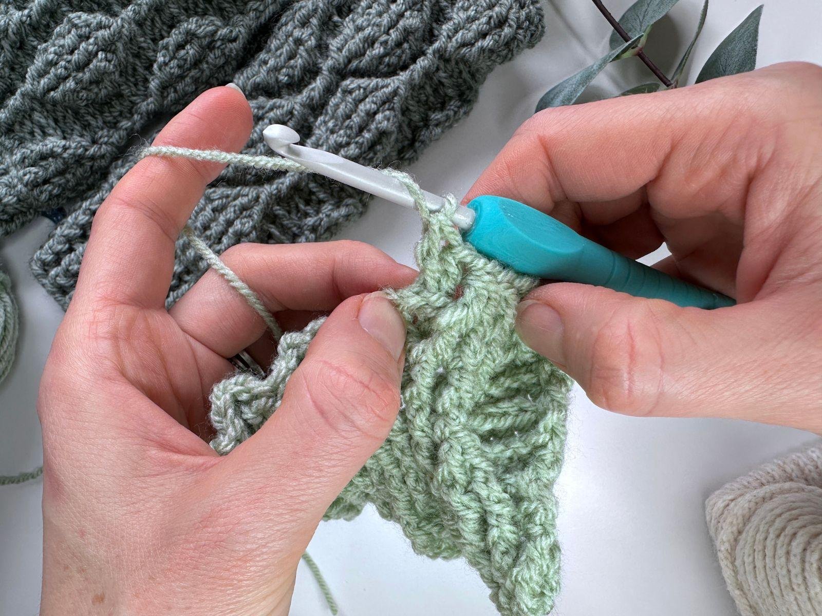 A crochet hook and 2 hands are working on a crochet fabric in green making post stitches in a pattern