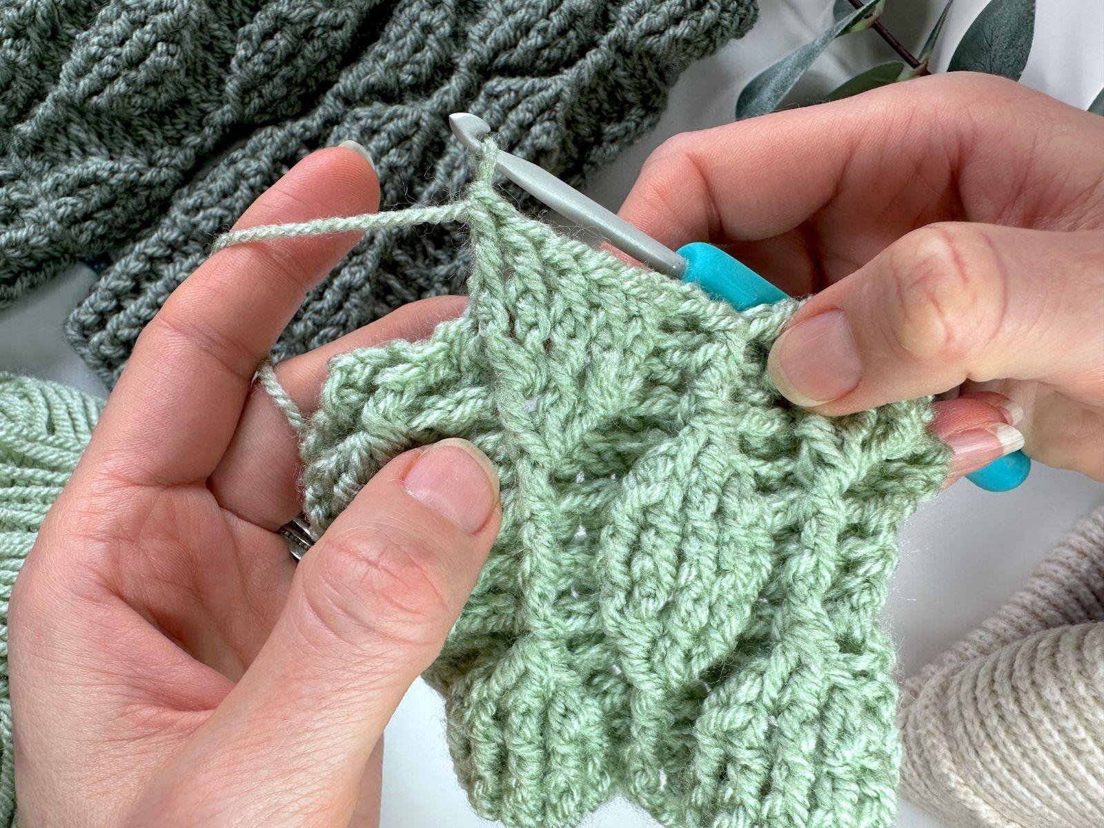 5 front post double crochet stitches in green yarn are made which create a leaf motif pattern. It is held in 2 hands along with a crochet hook