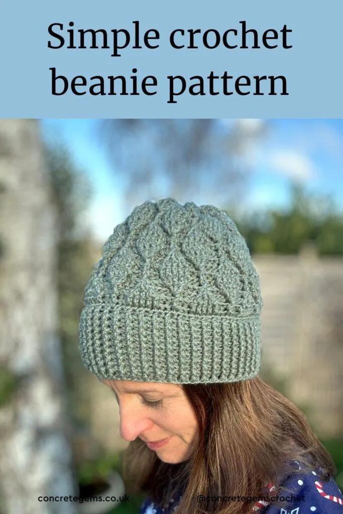 A lady stands sideways to the camera in a garden looking down wearing a green crochet beanie with textured crochet leaves running down the hat.