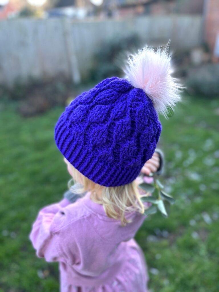 A small girl stands in a garden facing away from the camera. She wears a pink sweater dress and a purple crochet leaf hat with a pink pom pom.
